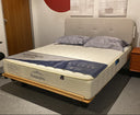 A twin-size mattress on a wooden platform bed frame with a light gray upholstered headboard, shown in a bedroom setting with an office chair, nightstand, and wall decor.